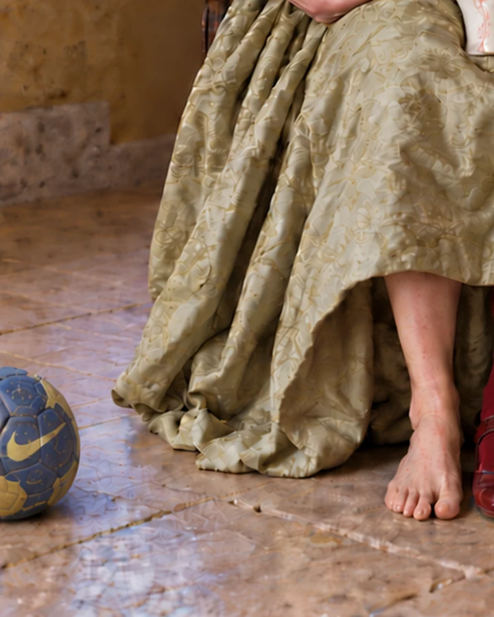 Detalle de la obra: un balón de fútbol gastado, azul y amarillo, queda parcialmente fuera del borde inferior izquierdo, junto al pie descalzo de una mujer adulta y los pliegues de su vestido renacentista verde.